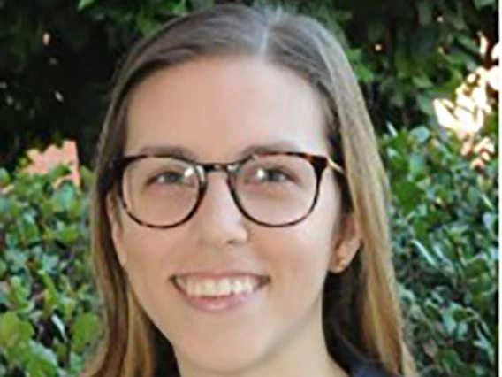 Smiling woman with long straight brown hair, wearing glasses, a navy blazer, and white lace top, standing outdoors with green foliage in background.
