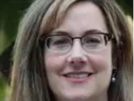 Woman with shoulder-length light brown hair and glasses, smiling and wearing a blue floral blouse and a large beaded necklace, with a blurred outdoor background.