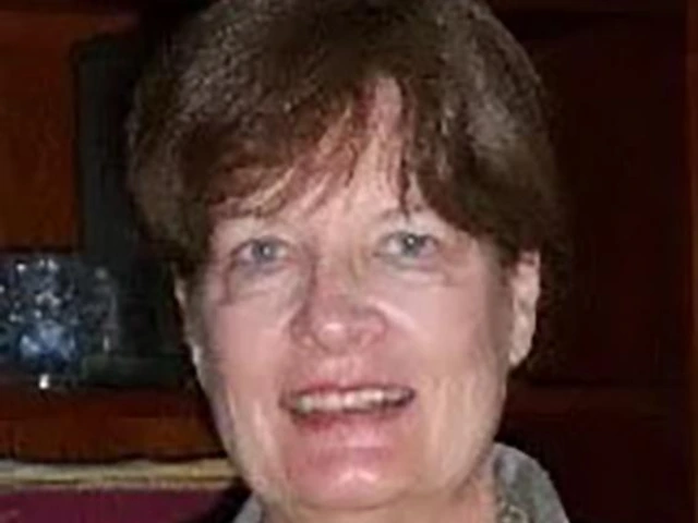 Smiling older woman with short brown hair, wearing a gray blazer and gold necklace, standing indoors in front of a wooden bookshelf.
