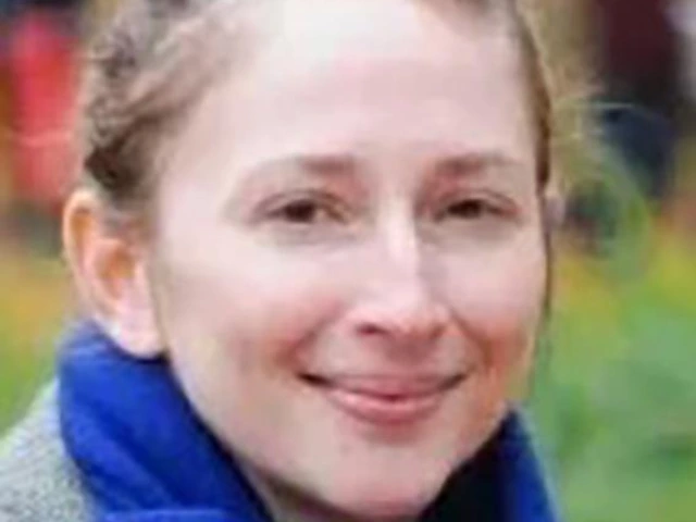 Woman with light brown hair pulled back in a bun, smiling slightly, wearing a gray coat and royal blue scarf, photographed outdoors with a soft-focus background.
