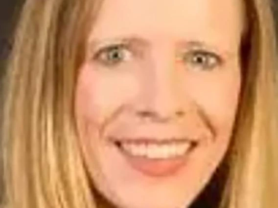 Smiling woman with long straight blonde hair, wearing a red jacket over a white top, gold necklace, and photographed against a dark background.