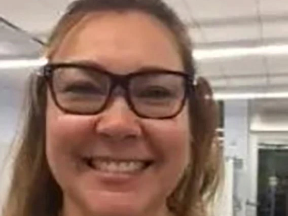 Smiling woman with light brown hair in a half-up style, wearing black-rimmed glasses and a gray shirt, photographed in a classroom with ceiling projectors.