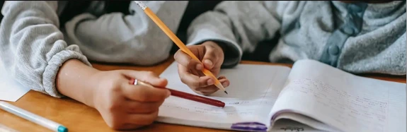 Close-up of two children working together at a desk, writing in an open notebook with pencils.