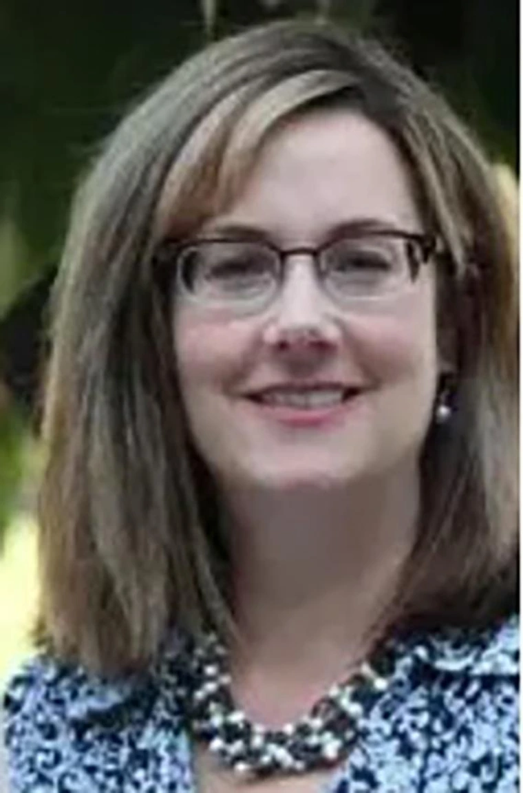 Woman with shoulder-length light brown hair and glasses, smiling and wearing a blue floral blouse and a large beaded necklace, with a blurred outdoor background.