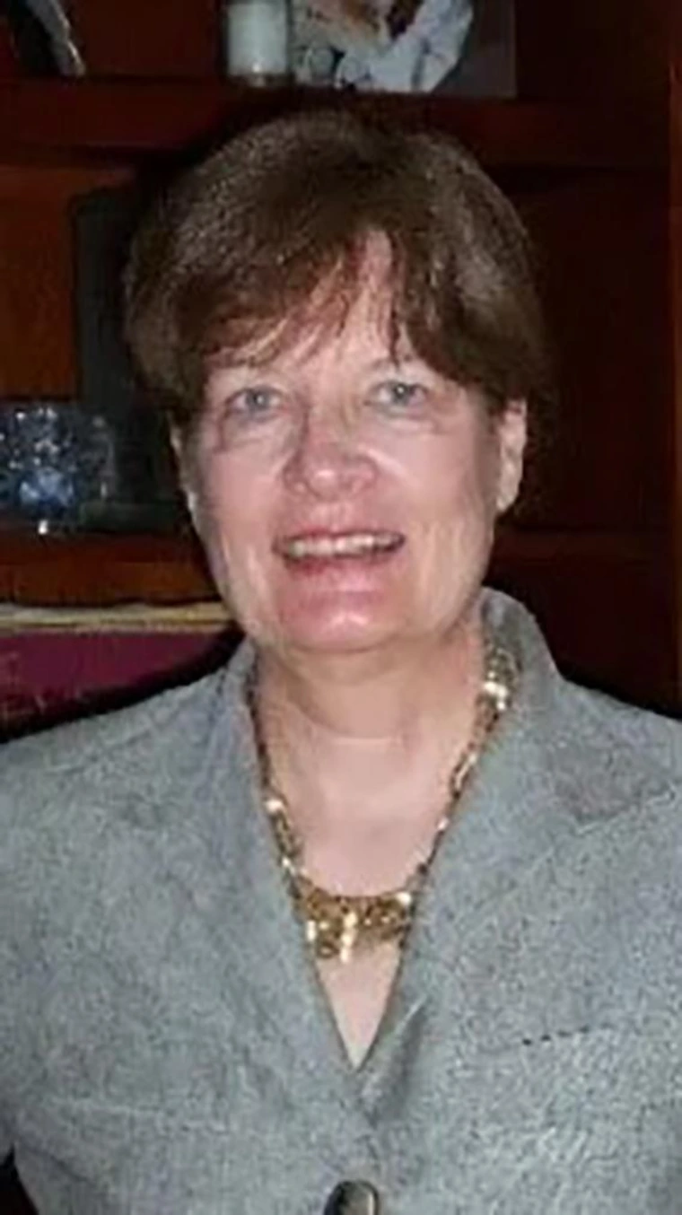 Smiling older woman with short brown hair, wearing a gray blazer and gold necklace, standing indoors in front of a wooden bookshelf.