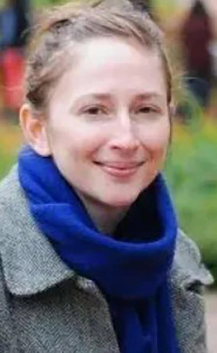 Woman with light brown hair pulled back in a bun, smiling slightly, wearing a gray coat and royal blue scarf, photographed outdoors with a soft-focus background.