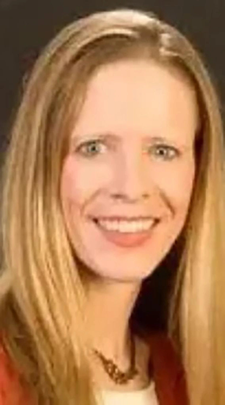 Smiling woman with long straight blonde hair, wearing a red jacket over a white top, gold necklace, and photographed against a dark background.
