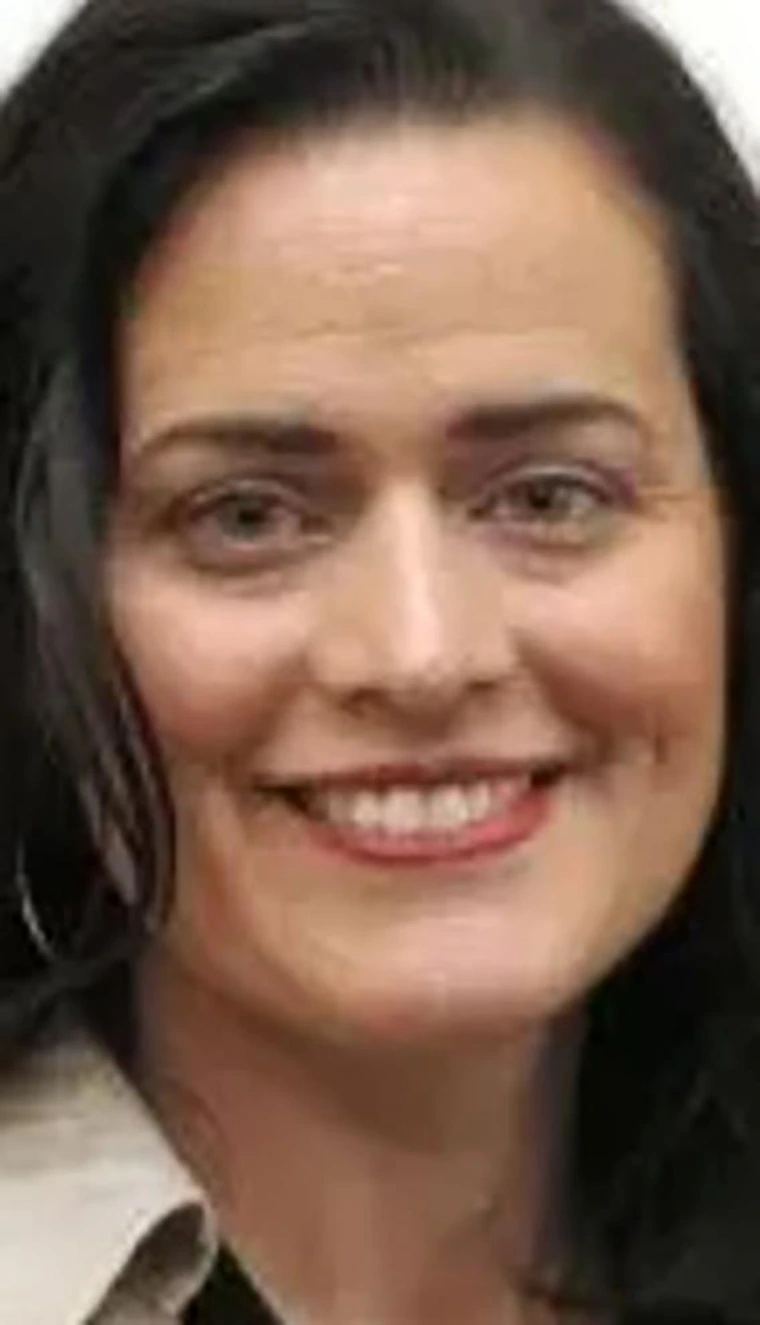 Smiling woman with long dark hair, wearing a light top and hoop earrings, photographed against a plain background.