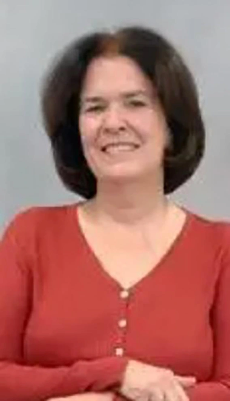 Smiling woman with short dark brown hair, wearing a red buttoned sweater, standing in front of a gray background.