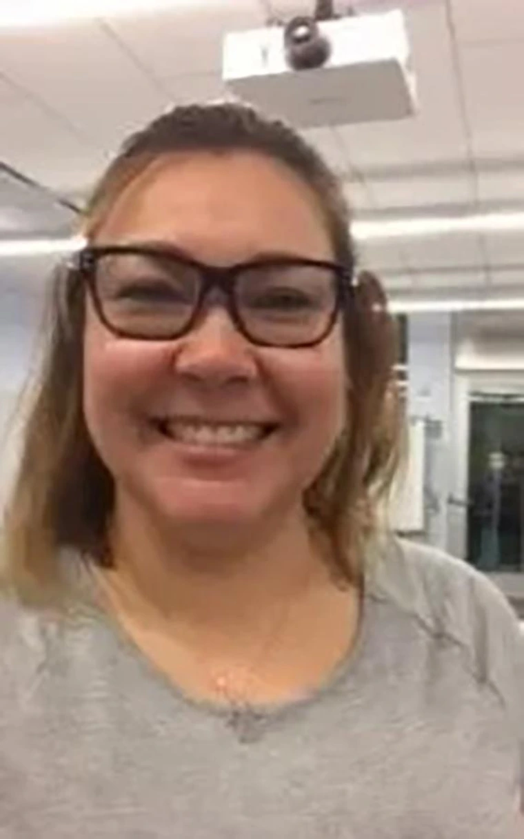 Smiling woman with light brown hair in a half-up style, wearing black-rimmed glasses and a gray shirt, photographed in a classroom with ceiling projectors.