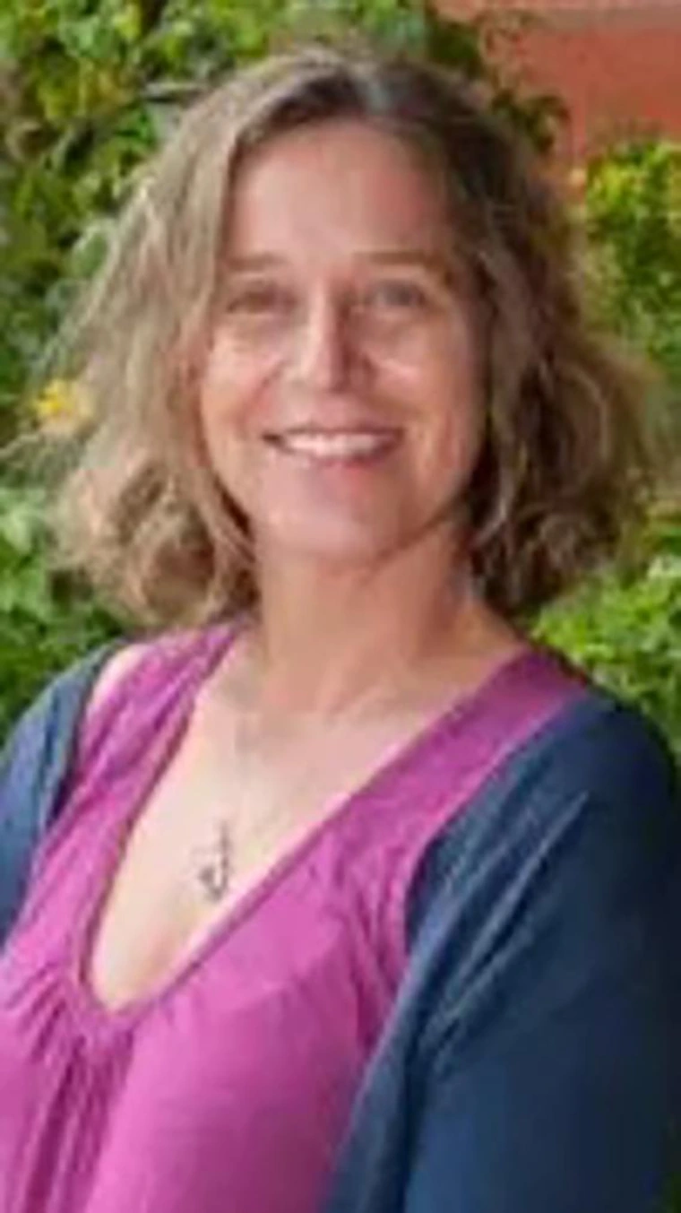 Smiling woman with shoulder-length blonde wavy hair, wearing a magenta blouse with a navy cardigan, photographed outdoors in front of green foliage.