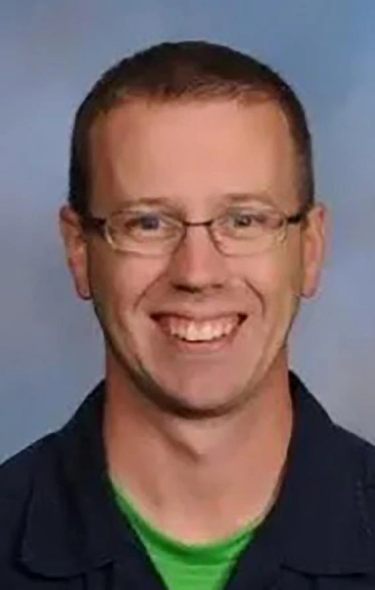 Smiling man with short light brown hair and glasses, wearing a dark jacket over a green shirt, with a neutral studio background.