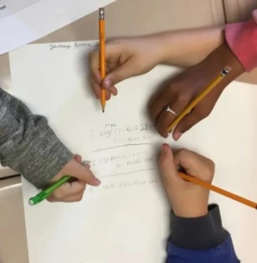 Four children's hands holding pencils collaboratively working on a math problem on white paper.