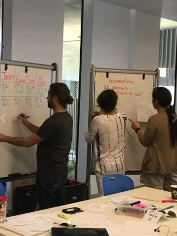 Three adults collaborating in a classroom or workshop setting, writing on large whiteboards with notes and diagrams, surrounded by tables and supplies.